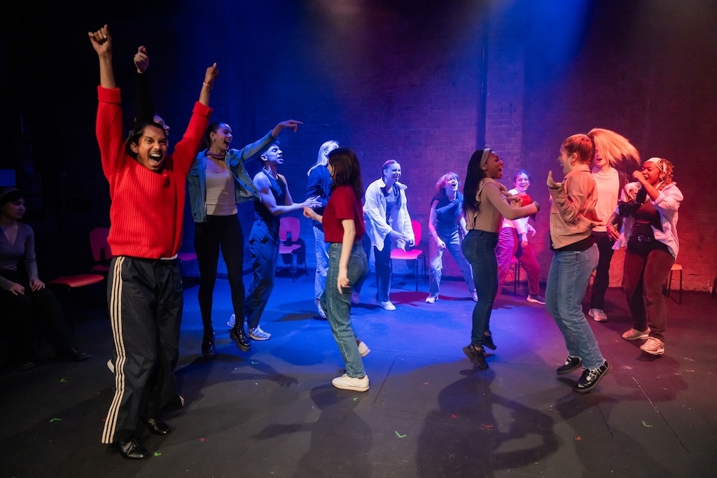 Action shot of the spring 2024 Shakespeare in Performance at RADA final presentation of Julius Caesar. Actors are in expressive mid-performance, some dancing, others jumping or shouting. They are all on stage under a blue/purple spotlight.